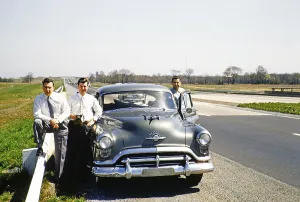 Three men in business suits standing next to an Oldsmobile 88 on a motorway, USA around 1953