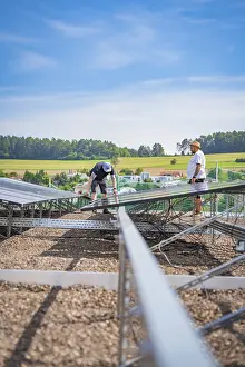Two men install solar panels on a construction site surrounded by rural fields