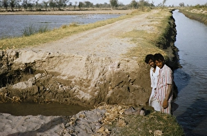 Two men stand on a broken country road next to a canal in rural Pakistan in 1962