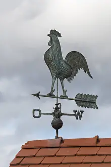 Metal weather vane with rooster on a tiled roof against cloudy sky