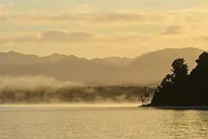 Mist drifts over a calm lake with mountains in the background at sunrise