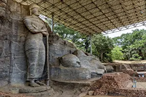 Monumental rock statue with standing and resting figure under a canopy surrounded by nature and brick walls