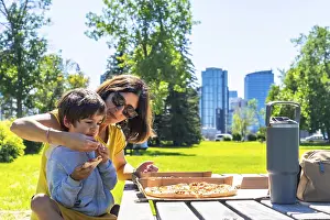 Mother helps her son eat a slice of pizza at a picnic table in a calgary park