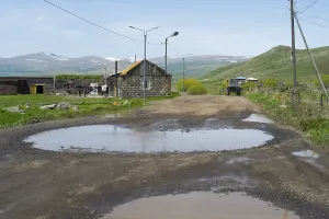 A muddy road with large puddles leads through a village with mountains in the background