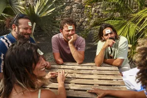 A multiethnic group of friends plays a guessing game at a wooden table outdoors They laugh while trying to identify