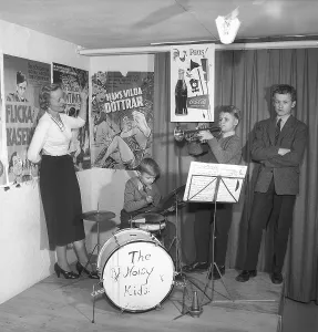 The Noisy Kids 1956. A drummer and a trumpeter are playing on a simple stage. On the wall behind them are film posters and advertisements for Coca-Cola. A woman stands to the side, listening