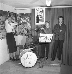 The Noisy Kids 1956. A drummer and a trumpeter are playing on a simple stage. On the wall behind them are film posters and advertisements for Coca-Cola
