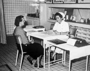 Old black and white archival photograph showing female factory worker visiting nurse at dispensary in the 1950s
