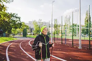 Old woman in sportswear practicing nordic walking outdoors on rubber treadmill in stadium
