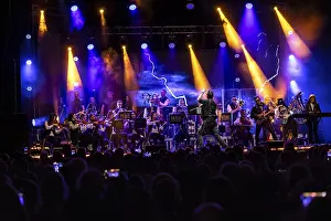 An orchestra plays on an illuminated stage in front of a large audience