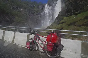 A packed bicycle stands on a road in front of an impressive waterfall in a mountainous landscape