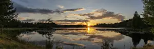 A panorama of a colorful sunset reflected in a calm lake landscape with green forest and reeds