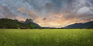 Panoramic view of a green meadow in front of imposing mountains under a cloudy evening sky