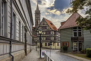 Framed Print : Paved street with half-timbered houses and church tower under a cloudy sky