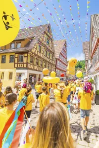 People in yellow clothes walk under colourful pennants in a narrow street in front of traditional houses