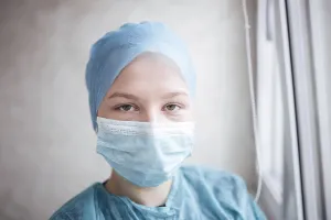 A person in blue scrubs and a mask looks reflective near a window