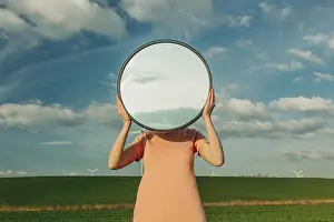 A person stands in a field, holding a circular mirror that reflects the blue sky and clouds