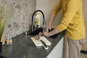 A person wearing a yellow shirt is washing utensils and a cutting board in a dark kitchen sink