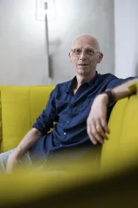 Portrait, older, bald man with glasses and blue shirt sits relaxed on a yellow sofa in a modern room
