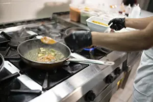 Professional chef wearing black gloves skillfully adding sauce to a meal preparation in a frying pan on the stove