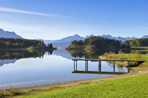 A quiet lake with a jetty, surrounded by trees and mountains under a clear blue sky