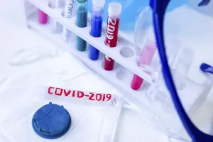 A rack with colored vials and a COVID-2019 test tube under safety glasses against a blue background