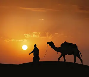 Rajasthan travel background, indian cameleer (camel driver) with camel silhouette in dunes of Thar desert on sunset