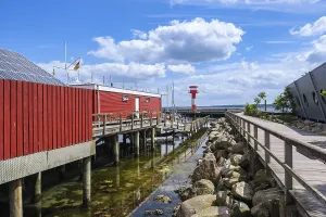 A red building on the waterfront with a jetty and rocks on the shore under a blue sky