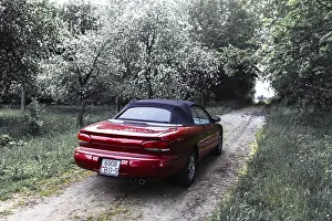 Red convertible car on a narrow path through dense forest with overcast skies