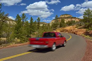 A red pickup truck drives on a winding road through a scenic mountain region with trees and blue sky