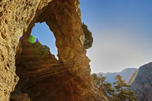 A rocky arch with sunbeams, surrounded by trees and mountains under a clear sky