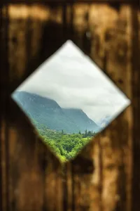 Rustic wooden frame reveals a lush green landscape with mountains in the background under a cloudy sky