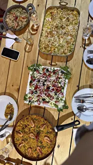 Rustic wooden table set with multiple homemade dishes including a large pan of seafood paella