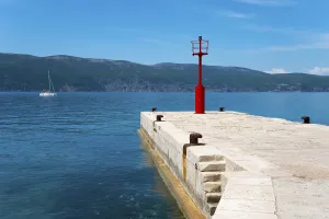 Sailing boat next to a stony jetty with a red lighthouse under a clear blue sky