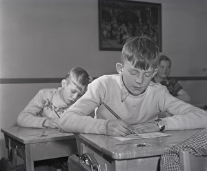 School in the 1940s. The interior of a classroom with a class of children sitting neatly at their desks. A boy is sitting at the front of the class, writing with an ink pen. The school desks had a lid that opened