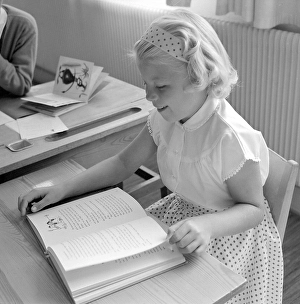 School in the 1950s. A girl is seen sitting at her desk in the classroom reading from the schoolbook in front of her