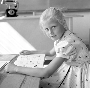 School in the 1950s. A girl is seen sitting at her desk in the classroom reading from the schoolbook in front of her