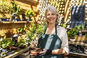 Senior gardener woman smiling while holding a small bonsai tree in a vibrant shop