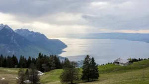A serene view of Lake Geneva surrounded by rolling alpine hills and trees Sunlight breaks through the clouds