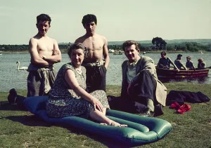 Two shirtless school boys kneel by mature woman possibly matron and young male teacher by side of rowing lake, England, UK 1950s
