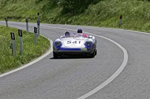 A silver racing car with two people drives swiftly along a winding road