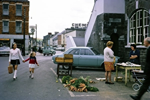 Small street market with carrots, people and traffic in Macroom, County Cork, Republic of Ireland, 1969