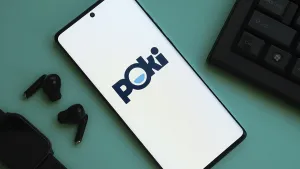 A smartphone displaying the Poki logo on a desk with earbuds and a keyboard