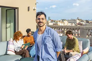 Smiling man standing on a rooftop terrace as diverse colleagues relax and work in the background