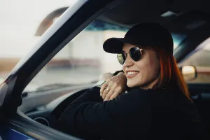 Smiling woman wearing sunglasses and a cap sits relaxed in a car