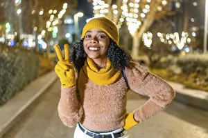 Smiling woman wearing yellow winter clothes showing victory sign while enjoying christmas lights in a city at night