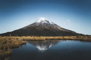 A snow-capped mountain is reflected in a still lake while people walk along the shore