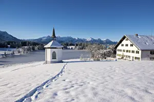 Snowy landscape with chapel and farmhouse against mountain backdrop under blue sky