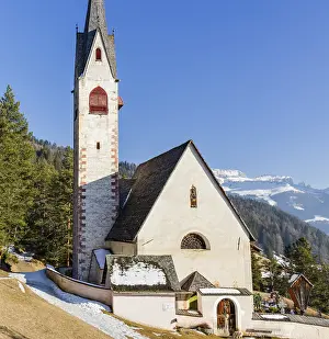 St. Jacob's Church overlooking pine forests and snow-capped peaks of Dolomites mountains in winter<br> St