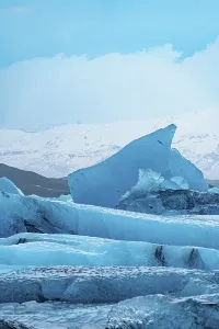 A stark blue iceberg rises against a cold wintry sky with snowy peaks in the distance